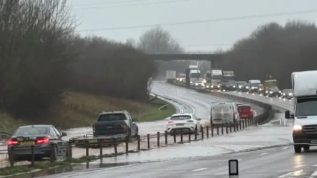 Inundaciones en la A30 cerca de Ottery St Mary. Hay coches y largos atascos en el lado izquierdo de la carretera, y coches inundados en el lado opuesto.