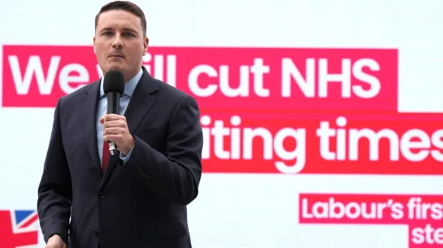 وزیر بهداشت بریتانیا میکروفون در دست دارد پشت او درباره بودجه ان هچ اس سیستم خدمات درمانی بریتانیا نوشته شده