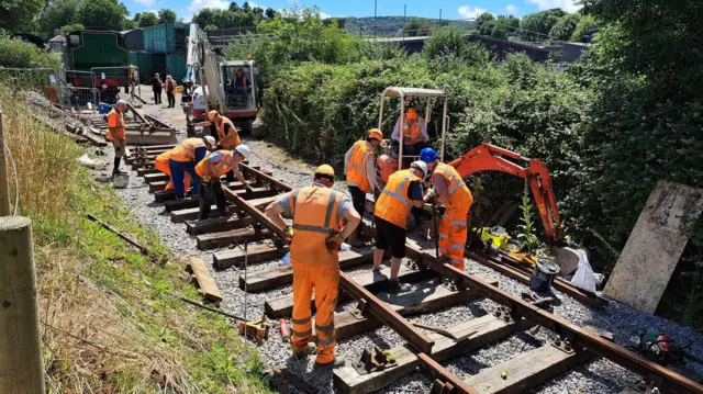 El fotógrafo se encuentra en un terraplén, observando a once personas con chalecos reflectantes naranjas y cascos de seguridad trabajando en una vía férrea en un día soleado. Dos miniexcavadoras se encuentran junto a la vía y los hombres colocan la última sección de acero sobre las traviesas. Al fondo, en la vía, se ve una locomotora de vapor verde.