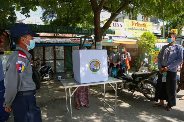 မဲပေးနေတဲ့အမျိုးသမီးနဲ့ ဘေးဘယ်ယာမှာရပ်နေတဲ့ အမျိုးသားတစ်ဦးနဲ့ ရဲတစ်ဦး 