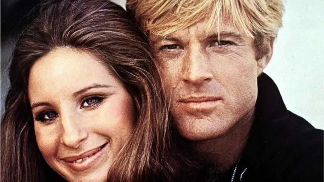Barbra Streisand et Robert Redford dans une photo promotionnelle tirée du film The Way We Were. Ils regardent tous deux droit vers l'appareil photo et Streisand sourit.