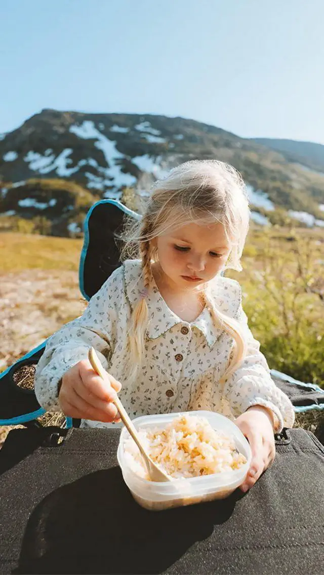 Menina come arroz em uma tigela plástica ao sol, em um campo aberto, com montanhas ao fundo