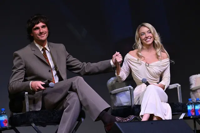 Елорді та Свіні сидять на сцені в кріслах, тримаючись за руки. Обидва тримають мікрофони; сцена затемнена, освітлення спрямоване на учасників, що створює атмосферу публічної розмови або фестивального заходу.