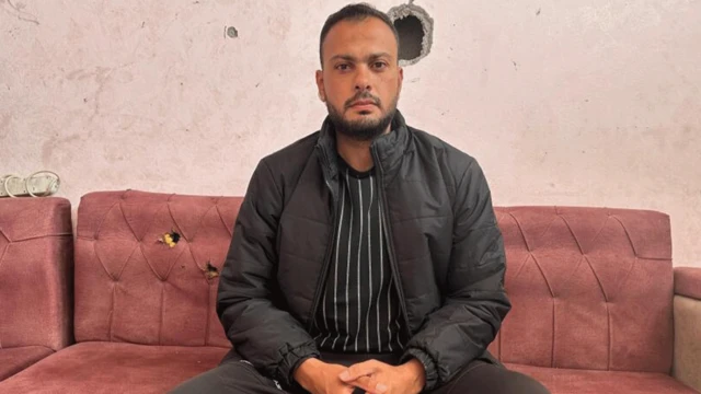 Moumen al-Natour, sentado em um sofá e olhando para a câmera. Ele veste uma camisa preta com listras verticais brancas, casaco e calças pretas. O sofá e a parede atrás dele têm buracos de bala.