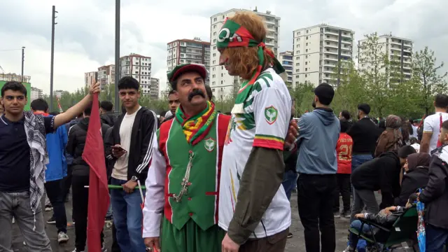 Mezher Tekçi ve Tommy yan yana ayakta dururken Tekçi, kendinden epey uzun olan Tommy'ye doğru yukarı bakıyor. Tekçi'nin büyük pos bıyıkları var. İkisinin de üstünde Amedspor formaları bulunuyor