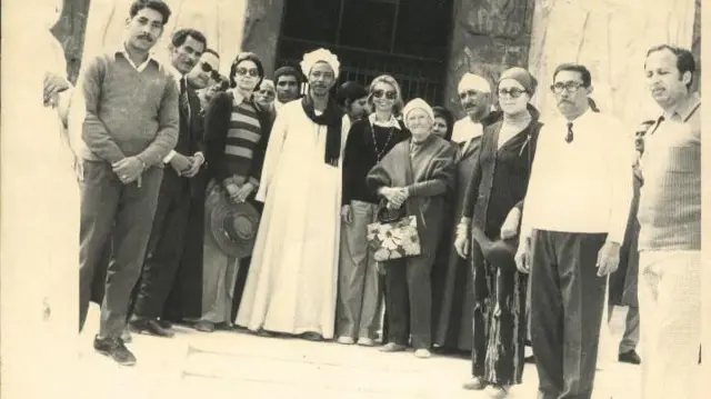 أم سيتي بوشاح أبيض على رأسها، تتوسط مجموعة من الزوار في أبيدوس. تحمل حقيبة عليها رسم لورود، على يسارها رجل بزي تقليدي مصري وعلى يمينها ارمأة تلبس نظارة سوداء.