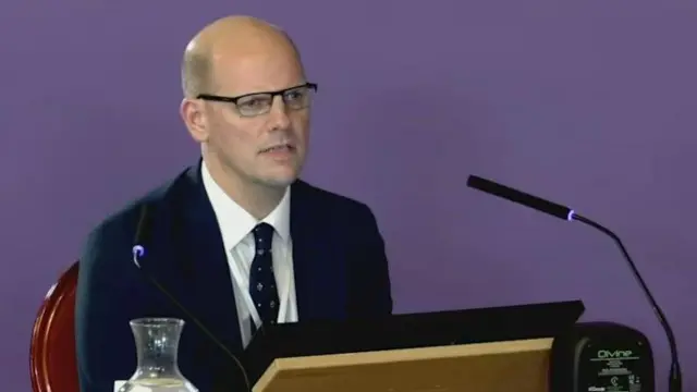 El Dr. Anthony Molyneux, que es calvo y usa gafas con marco negro, está sentado en un escritorio frente a una pantalla de computadora y micrófonos con una pantalla violeta de fondo.