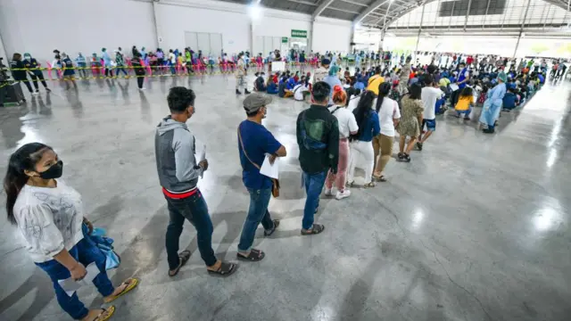 ထိုင်းမှာ ကိုဗစ်ကာလ ကာကွယ်ဆေးထိုးဖို့ တန်းစီစောင့်နေတဲ့ မြန်မာလုပ်သားတွေ၊ အမျိုးသားအမျိုးသမီးတွေ အဆောက်အဦးကျယ် တစ်ခုအောက် တန်းစီပြီး မတ်တပ်ရပ်နေကြ။