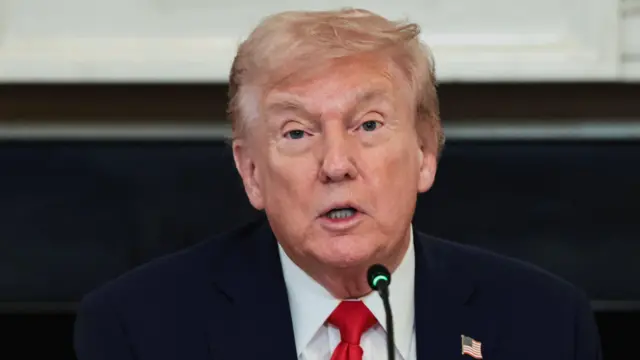 Primer plano de Trump ante un micrófono. Está vestido con traje oscuro, camisa blanca y corbata roja. Tiene en la solapa un pin con la bandera de Estados Unidos.