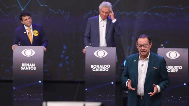 Em um palco de debate, três candidatos aparecem diante de púlpitos identificados com seus nomes. À esquerda, Renan Santos observa a cena; ao centro, Ronaldo Caiado leva a mão ao ouvido; e, em primeiro plano, Augusto Cury fala gesticulando. O fundo exibe gráficos em tons azulados e elementos visuais do evento.