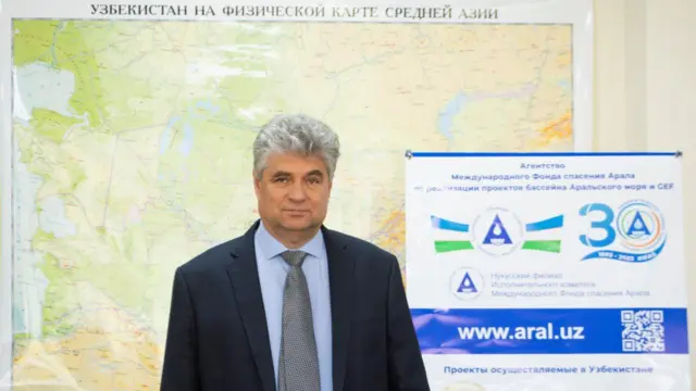 Rasm markazida Vadim Sokolov, uning orqa fonida o‘ngda egntlikning baneri, umuniy orqa fonda esa Makraziy Osiyoning tabiiy geografik xaritasi