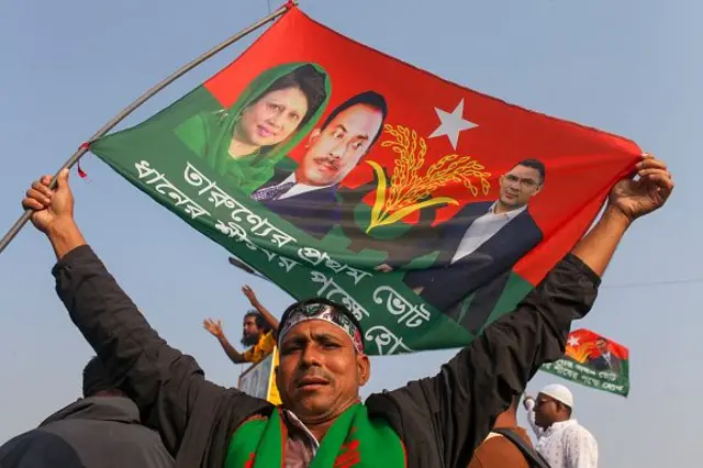 এক ব্যক্তি মাথার উপর দুই হাত মেলে পতাকা তুলে রেখেছে যেখানে জিয়াউর রহমান, খালেদা জিয়া, তারেক রহমান এবং ধানের শীষের ছবি রয়েছে। নীচে লেখা 