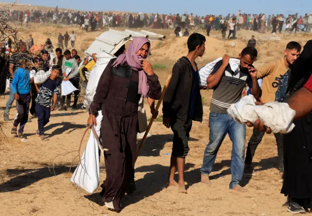Bir kadın elinde çuvalla yürüyor. Arkasında çuvallar taşıyan çok sayıda erkek var.