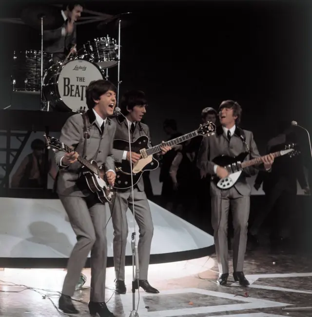 Paul McCartney toca la guitarra y canta frente a un micrófono. A su lado está George Harrison, también con su guitarra. A la izquierda está John Lennon. Atrás está la batería que dice The Beatles. Los cuatro miembros están vestidos igual con camisa blanca, corbata negra y chaqueta y pantalón grises. Están en una presentación
