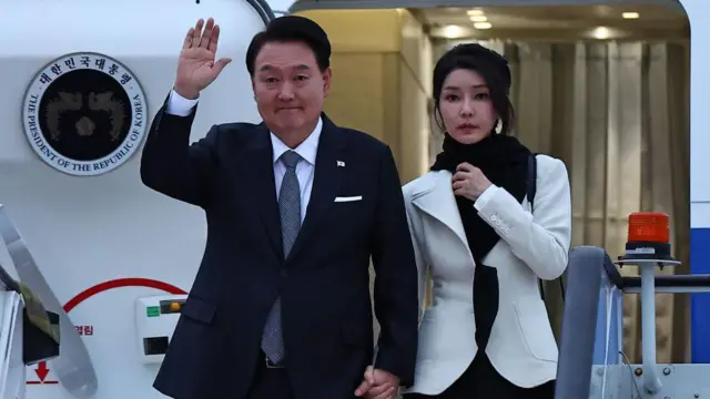 Kim Keon Hee and Yoon Suk Yeol saindo de um avião, de mãos dadas. Ele acena para as câmeras.