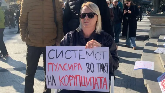 Žena u invalidskim kolicima drži transparent sa natpisom - Sistem pulsira u ritmu korupcije