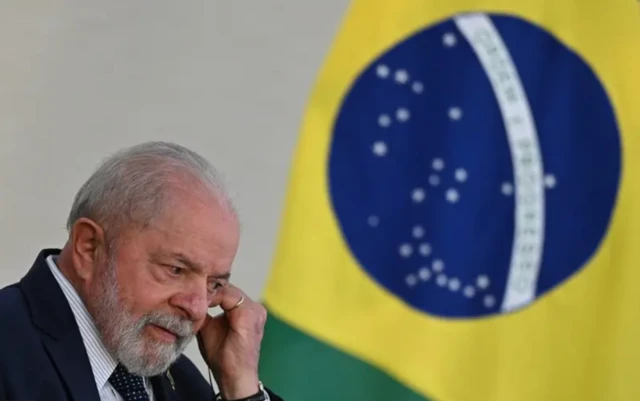 O presidente Luiz Inácio Lula da Silva posicionando um fone de ouvido com uma bandeira do Brasil ao fundo