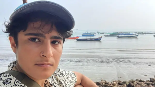 Sophia sentada na praia para uma selfie, de camisa estampada, boné de baseball e barcos de pesca ao fundo