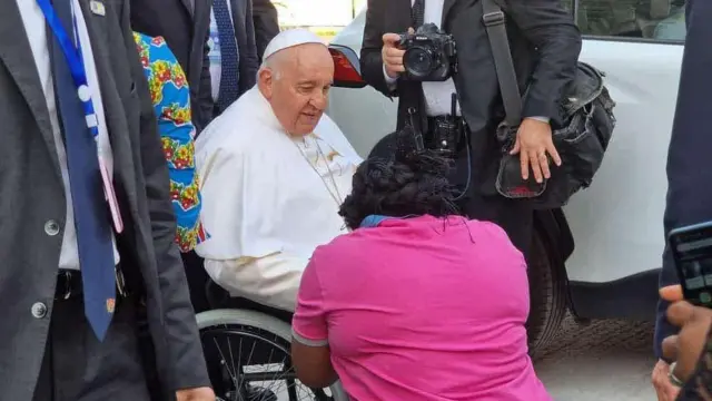 Le pape François, vêtu d'habits blancs, est assis sur un fauteuil roulant et regarde Kitsita Ndongo Rachel, qui porte un haut rose et s'est agenouillée. Autour d'eux se tiennent des personnes vêtues de costumes gris. L'une d'entre elles porte un sac d'appareil photo noir et tient un appareil photo Nikon noir.