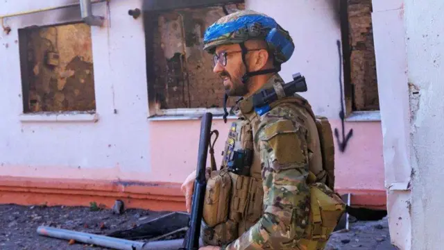 Umusirikare wa Ukraine afite imbunda ya Kalashnikov, arimo kugenda n'amaguru anyuze iruhande rw'inyubako yarashwe amadirishya akavamo. Ifoto yafashwe muri Nzeri (9) mu mwaka wa 2024, mu gace ka Sudzha, mu karere ka Kursk, mu Burusiya. 


