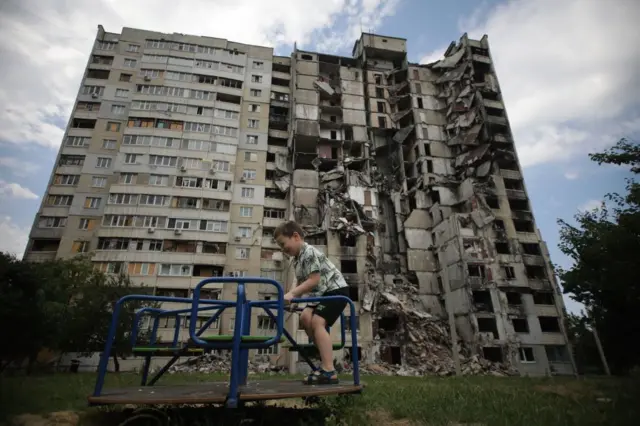 Хлопчик катається на каруселі на подвір’ї житлового масиву. Позаду височіє багатоповерховий будинок, значна частина якого повністю зруйнована: секція посередині обвалилася, оголивши квартири та уламки перекриттів. Навколо — зелена трава та дерева, а небо вкрите легкими хмарами. 