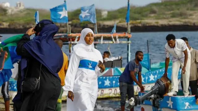 امرأة ترتدي حجاباً أبيض وفستاناً  يمزج بين اللونين الأبيض والأزرق من أعلام الصومال، بينما  تسير بجوار القوارب الراسية على شاطئ البحر.