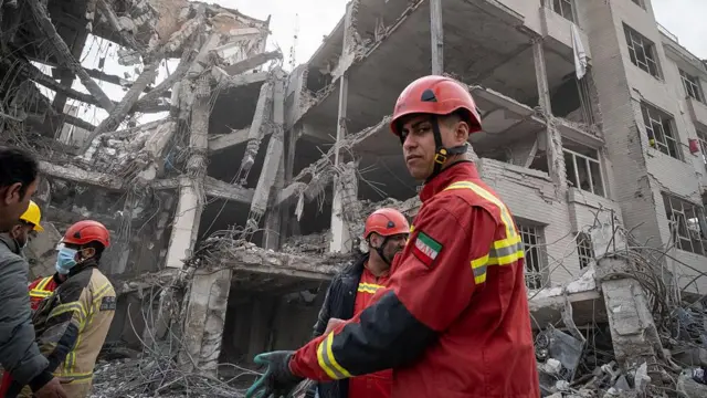 یک آتش‌نشان ایرانی روی خرابه‌های ساختمان‌هایی که در جریان عملیات نظامی آمریکا و اسرائیل به یک منطقه مسکونی در تاریخ ۹ مارس آسیب دیده‌اند، ایستاده است – تهران، ایران، ۱۲ مارس ۲۰۲۶.