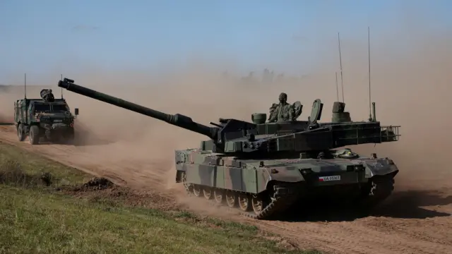 Czołg jedzie ciemną, zakurzoną drogą, obok niego transporter opancerzony. W wieżyczce czołgu stoi żołnierz.