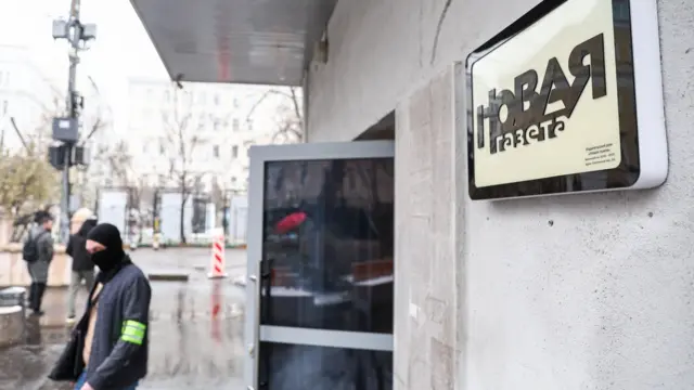 Здание с логотипом Новой газеты, перед открытой дверью своит мужчина в балаклаве.