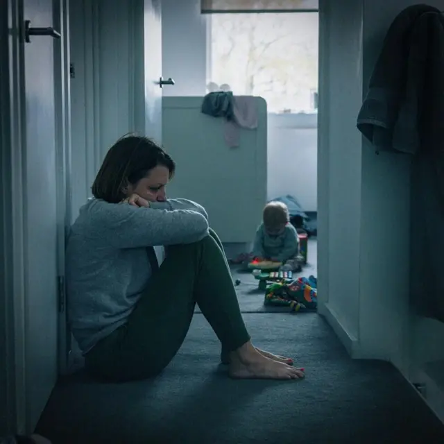 Una madre sentada en el piso cabizbaja. En el fondo de ve a un niño pequeño jugando.