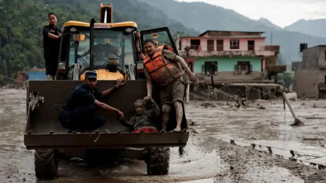 Rescuers use one digger truck to carry an elderly woman to safety. Buildings swamped by mud can be seen in the background
