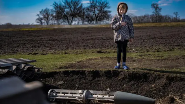 Даша, 8 років, стоїть на сільськогосподарському полі біля снаряда, якого ніхто не хоче торкатися; селяни чекають саперів ДСНС для вилучення снаряда 1 березня 2023 року, Трудолюбівка, Херсонська область.
