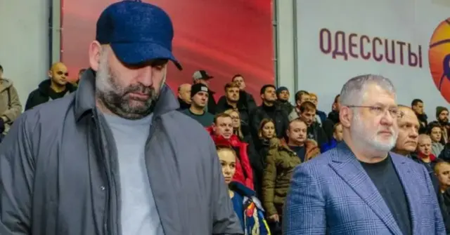 Міндіч в сірій куртці і синій кепці дивиться вниз. Поруч з ним Коломойський в синьому піджаку дивиться прямо