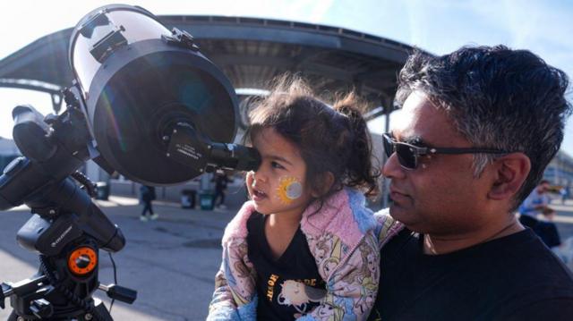 Homem de pele morena segura criança de pele clara com sol e lua desenhados no rosto com o rosto encontrado no visor de um grande telescópio