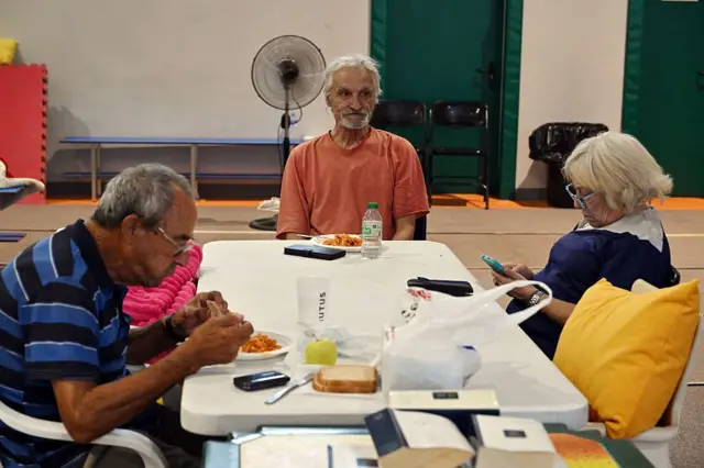 Tres personas sentadas alrededor de una mesa de plástico comen y miran el teléfono dentro de un centro de refugio improvisado. 