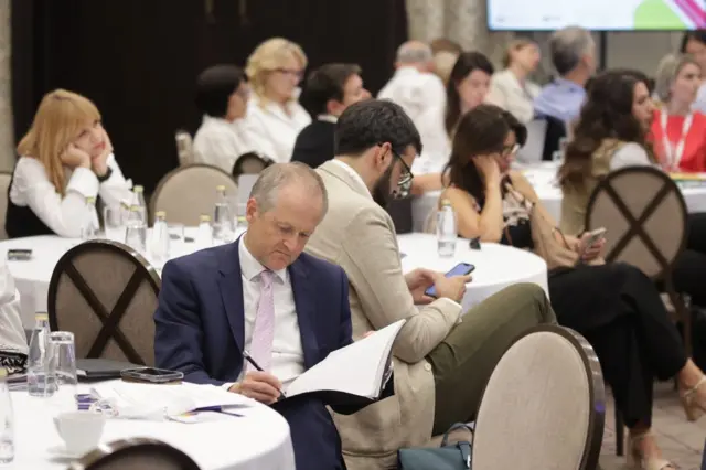 Vilijam Midlton u teget odelu sa roze kravatom sedi za stolom i čita