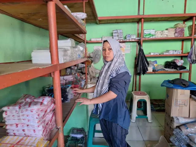 Hastina, dengan hijab dan baju lengan panjang biru sedang berdiri berhadapan dengan etalase warung yang berisi dengan tumpukan pak plastik. 