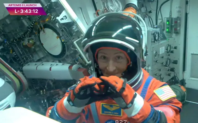 Bir astronot kadın kameraya kalp işareti yapıyor. Üzerinde turuncu uzay kıyafeti var