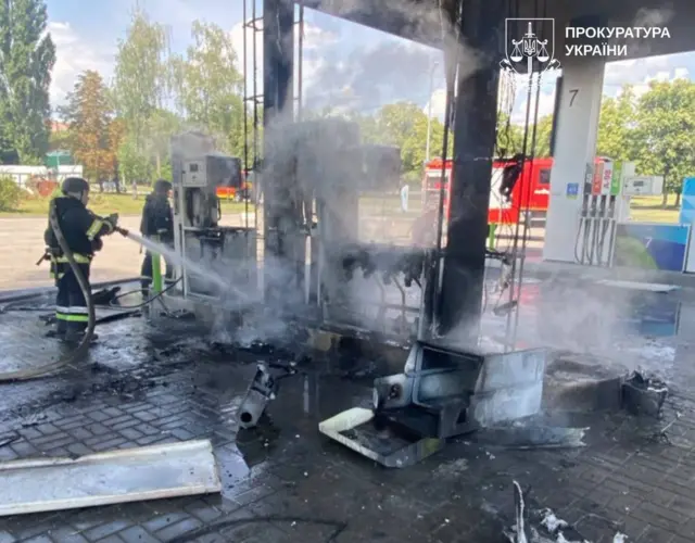 Пошкоджена АЗС після пожежі. Два пожежники гасять згорілу колонку, з якої ще підіймається густий дим. Навколо лежать обгорілі уламки конструкцій, а на задньому плані видно червоний автомобіль ДСНС.