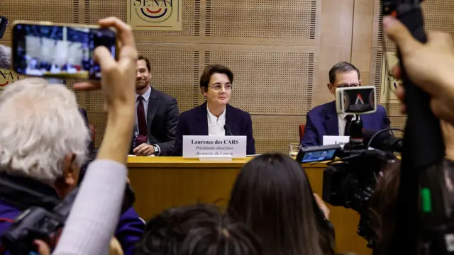 Cámaras de celular toman imágenes de la directora del Louvre sentada ante un escritorio y acompañada de dos hombres