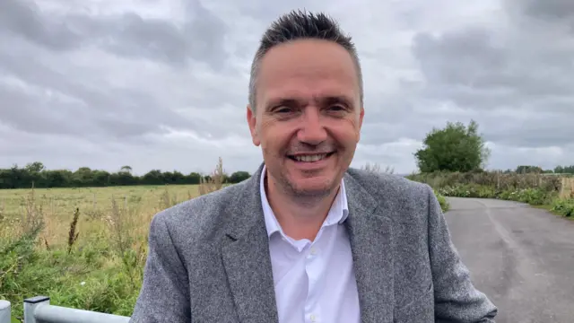 Mike Bell. Tiene el pelo corto y gris, y barba incipiente. Aparece en la foto junto a una carretera y un campo, con camisa blanca y blazer gris. Mira directamente a la cámara y sonríe.