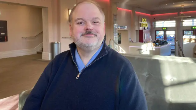 Rob Salmon, director de participación creativa, está sentado en un sofá gris en el vestíbulo del Teatro Stephen Joseph. Tiene el pelo corto y canoso, barba corta y viste un jersey y una camisa azules.
