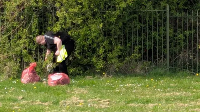 Paul Fairbridge collecting rubbish in a field. he is wearing a black shirt and a hi-vis jacket. He is bending down to collect rubbish. He has filled two red rubbish bags.
