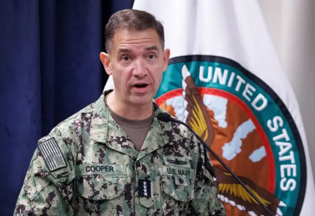 迷彩服を着たクーパー司令官が記者会見場らしき場所で話している