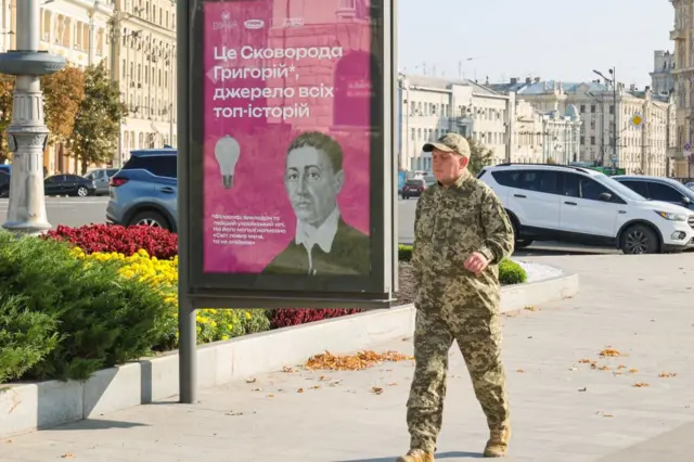 Сітілайт-постер із яскравим рожевим фоном і текстом українською:
"Це Сковорода Григорій, джерело всіх топ-історій". Нижче дрібніший текст: "філософ, викладач та перший український хіпі". Сітілайт стоїть на тротуарі, поряд із клумбами з жовтими та червоними квітами, повз йде чоловік у військовій формі. На задньому плані – кілька автомобілів, старі світлі будівлі та широка вулиця.