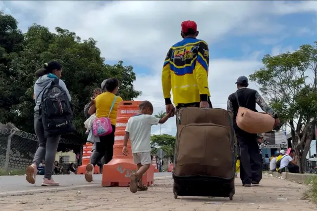 Grupo de refugiados caminha após cruzar a fronteira entre Venezuela e Brasil com malas de viagem e árvores ao fundo no cenário