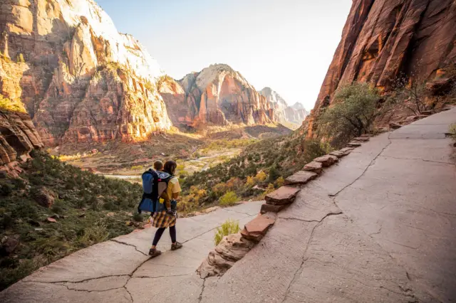 Una mujer camina por el parque Zión cargando a su bebé y al fondo se ven unas enormes montañas.
