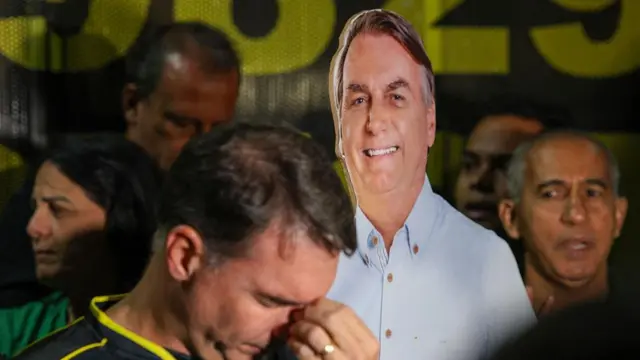 Flávio Bolsonaro com a cabeça baixa na frente de uma imagem de Bolsonaro em tamanho real.