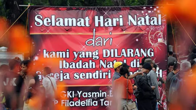 Sekelompok orang berdiri di sekitar spanduk raksasa bertuliskan "Selamat Natal dari kami yang dilarang Ibadah Natal di gereja sendiri".