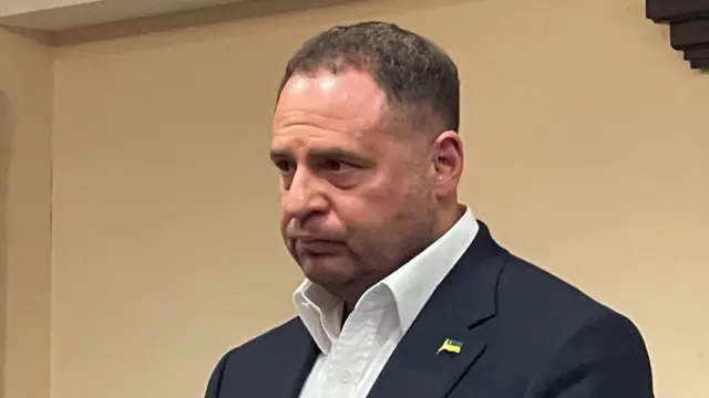 Єрмак крупним планом у темному піджаку та білій сорочці, на лацкані видно маленький значок із прапорцем. Голова злегка нахилена вперед, губи щільно стиснуті. Обличчя напружене.